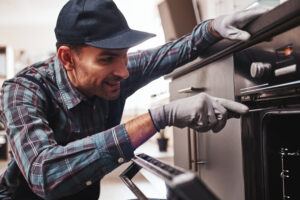 Appliance repairman examining oven with screwdriver in kitchen with tool case
