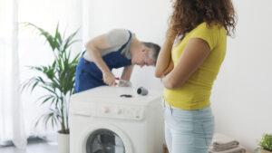 Appliance repairman repairing a broken washing machine, the customer is looking at him