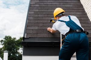 Handyman in uniform and helmet repairing roof while standing on ladder